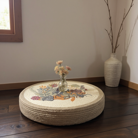 Round woven ottoman with floral design on a wooden floor, next to a vase with flowers and a tall vase.