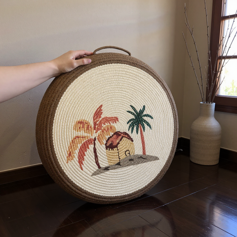 Round woven mat with palm tree and hut design held by a hand on a wooden floor.