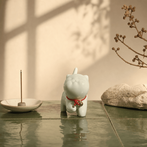 White ceramic cat figurine with a red collar on a reflective surface, with an incense holder and plant in the background.