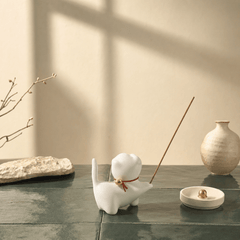 White ceramic dog-shaped incense holder with a lit incense stick on a dark surface.