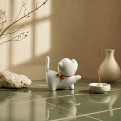 White ceramic cat figurine with a bell on a reflective surface, with a vase and branch in the background.
