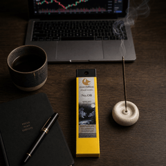 Incense stick holder with incense stick, yellow box, notebook, pen, and cup on a wooden surface.