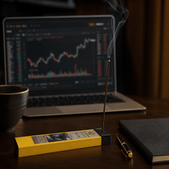 Laptop displaying stock market graphs on a desk with a lit incense stick and notebook.