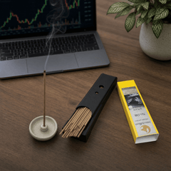 Incense stick holder with burning incense, black incense box, and yellow box on a wooden table with a laptop in the background.