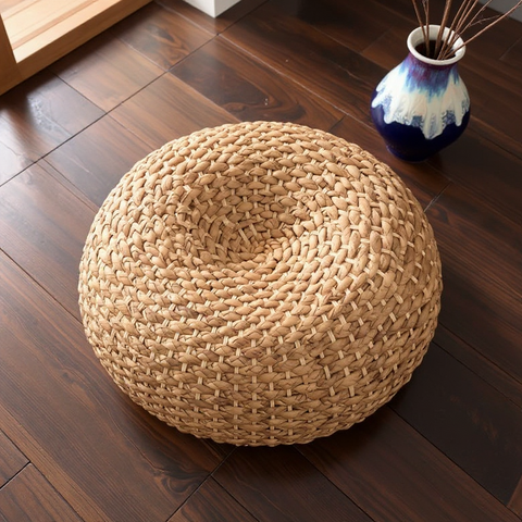 Woven beige ottoman on a wooden floor with a decorative vase in the background.