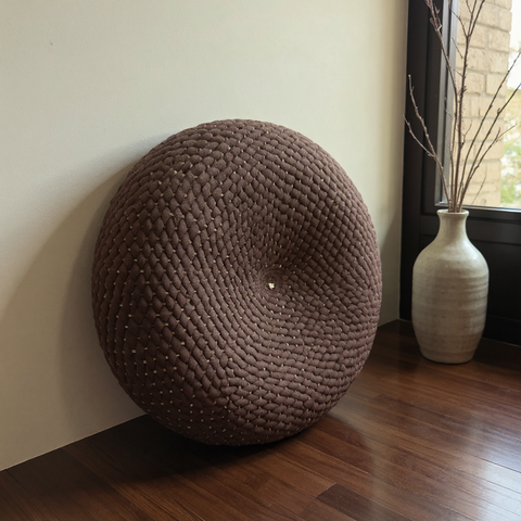 Round woven brown cushion on a wooden floor next to a vase with branches.